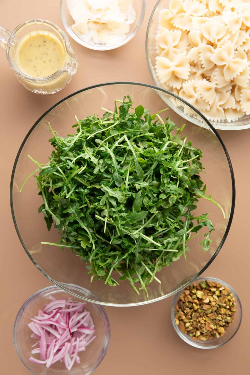pasta salad ingredients: lemon dressing, parmesan cheese, bowtie pasta, arugula, red onions, chopped pistachio nuts