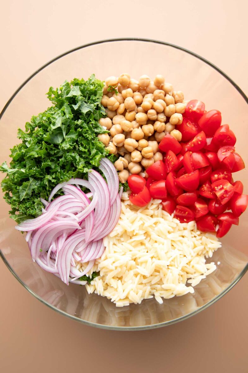 glass mixing bowl with chopped kale, chickpeas, cooked orzo, quartered cherry tomatoes, sliced red onion, and chickpeas