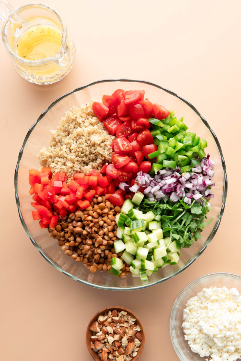 glass mixing bowl with quinoa, green bell pepper, cherry tomatoes, red bell pepper, lentils, cucumber, arugula and small topping bowls with feta, dressing, and almonds