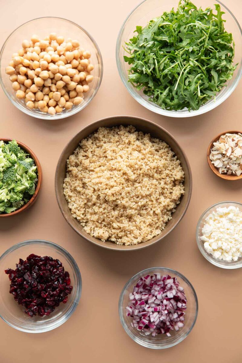 mixing bowls with salad ingredients: cooked quinoa, chickpeas, arugula, chopped broccoli, sliced almonds, crumbled feta cheese, dried cranberries, and chopped red onions