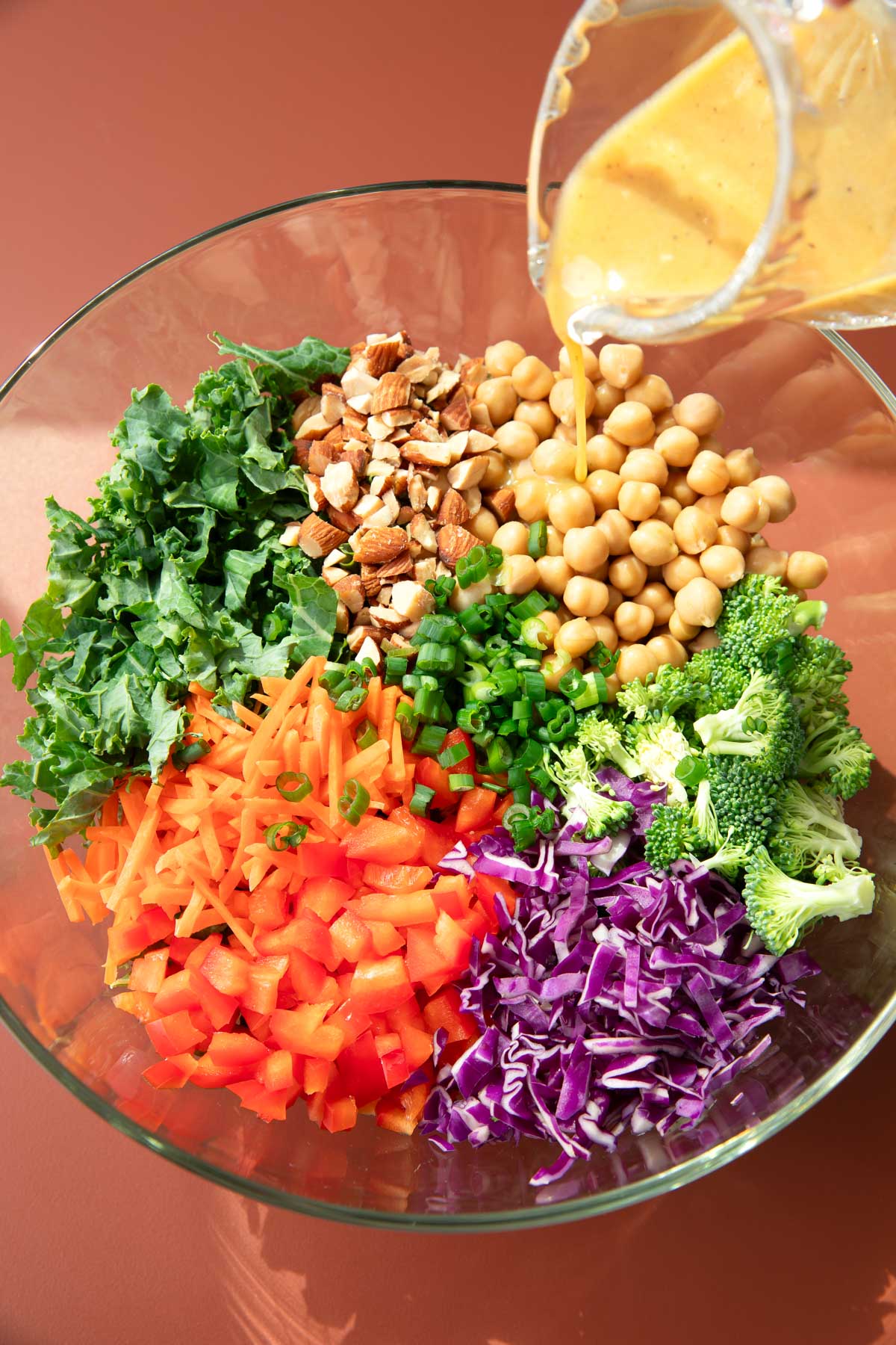 mixing bowl with kale, chickpeas, chopped almonds, shredded carrots, chopped bell pepper, shredded purple cabbage, chopped broccoli, and sliced green onions with dressing being poured on top